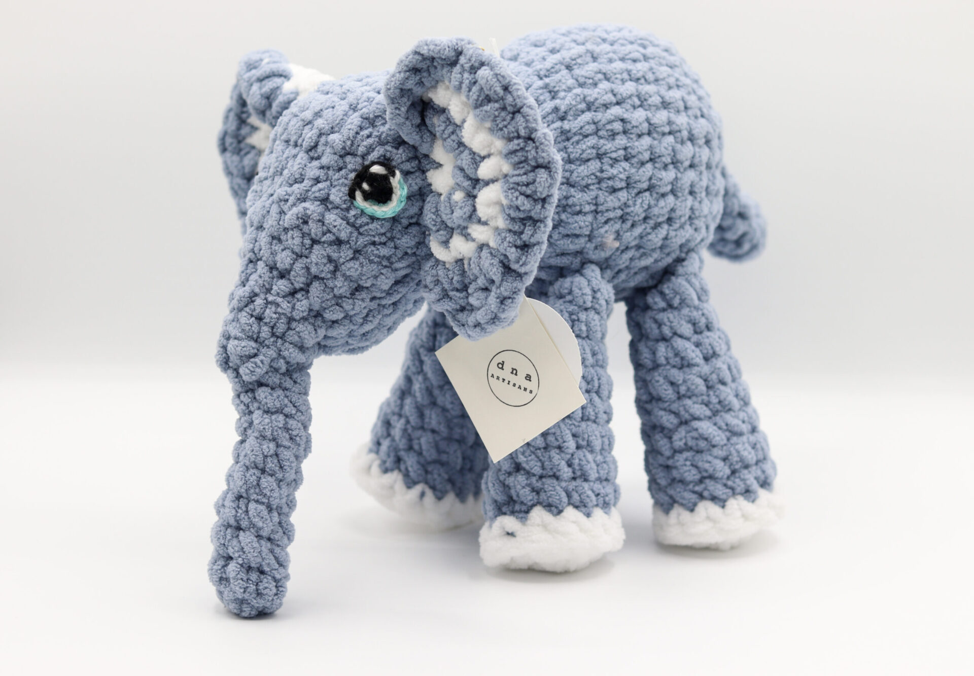 Photo d'une peluche éléphant tricotée à la main en laine bleue avec des détails blancs et un petit œil expressif. Cette peluche est douce et idéale pour les enfants.