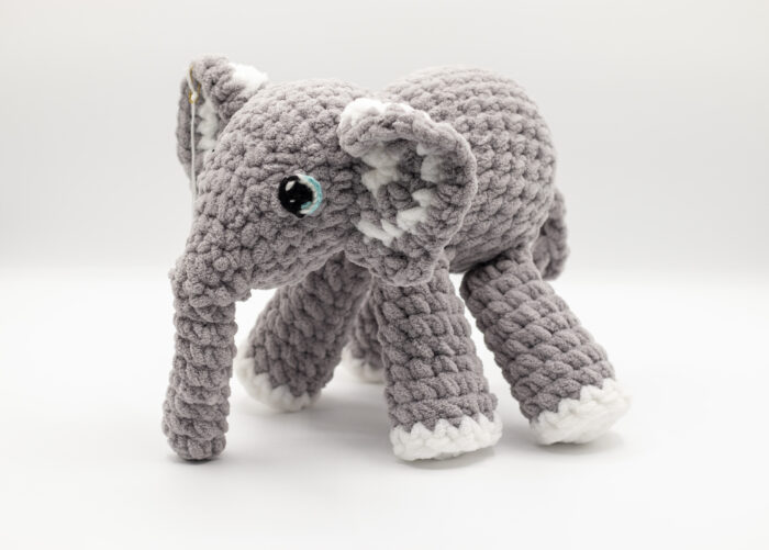 Photo d'une peluche éléphant tricotée à la main en laine grise et blanche avec des détails aux yeux. Cette peluche est douce et idéale pour les enfants.