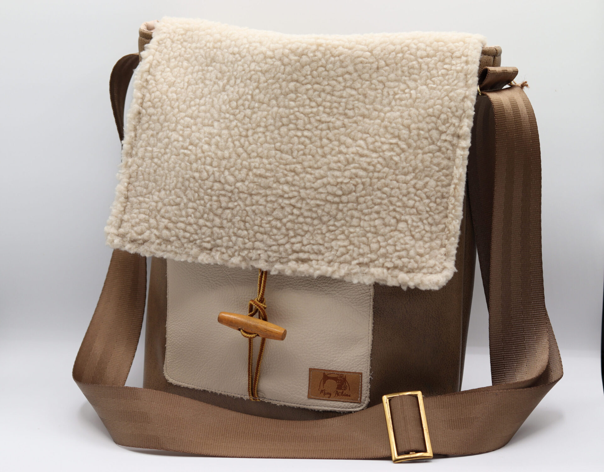 Photo d'un sac à main éco-responsable avec un rabat en tissu effet mouton blanc, une sangle ajustable marron et des détails en cuir beige et brun. Ce sac combine style rustique et modernité.