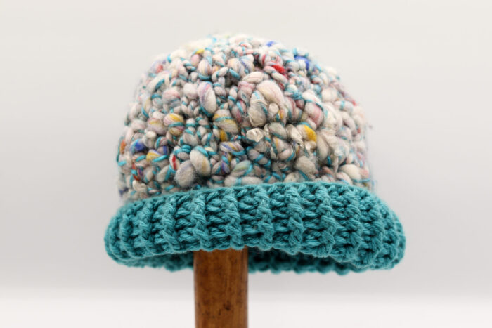 Bonnet artisanal en laine bouclée turquoise, aux nuances pastel de bleu, rose, violet et blanc, avec des touches discrètes de jaune et rouge. Le bord est crocheté en maille côtelée d'un bleu turquoise vibrant, offrant une finition éclatante et moderne. Ce bonnet unique, aux couleurs douces et à la texture moelleuse, est parfait pour apporter une touche de style tout en restant bien au chaud.