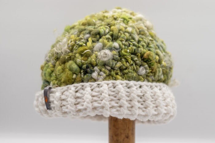 Bonnet en laine bouclée dans des nuances de vert mousse, jaune et blanc, avec une texture riche et naturelle. Le bord du bonnet est crocheté en maille épaisse d'un blanc cassé, apportant une touche douce et lumineuse. Un petit détail métallique orné d'un motif orange est visible sur le côté, ajoutant une note discrète et élégante à cet accessoire artisanal parfait pour les saisons fraîches.