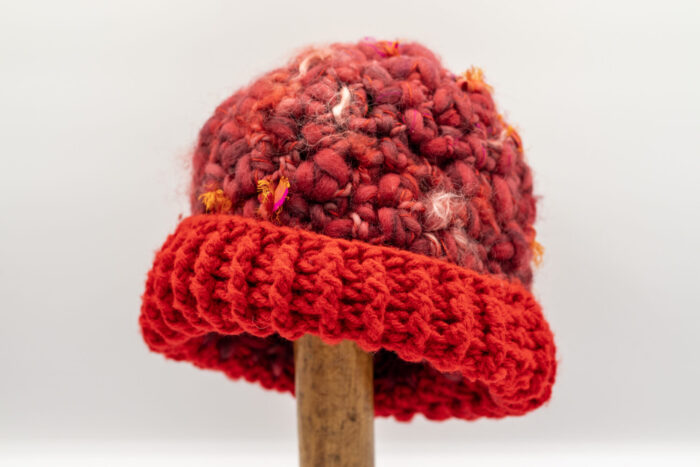 Bonnet en laine bouclée dans des nuances de rouge profond et bordeaux, agrémenté de touches colorées jaunes et roses. Le bord est crocheté en maille épaisse d'un rouge éclatant, offrant une allure chaleureuse et audacieuse. Cet accessoire artisanal allie confort et originalité, parfait pour les journées fraîches.