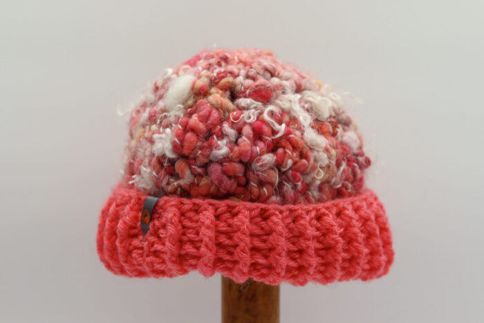 Bonnet artisanal en laine bouclée dans des tons de rose, rouge et blanc, offrant une texture douce et duveteuse. Le bord du bonnet est crocheté en maille côtelée d'un rose vif, créant un contraste éclatant. Un petit détail métallique avec un motif orange est visible sur le côté, ajoutant une touche unique à cet accessoire chaleureux et fait main.