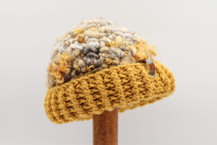 Bonnet artisanal en laine bouclée, avec une palette de couleurs beige, gris et jaune moutarde. Le bord du bonnet est crocheté en maille côtelée d'un jaune vif, ajoutant une touche chaleureuse et vibrante. Un petit détail métallique avec un motif orange est attaché sur le côté, renforçant le style unique et fait main de l'accessoire.